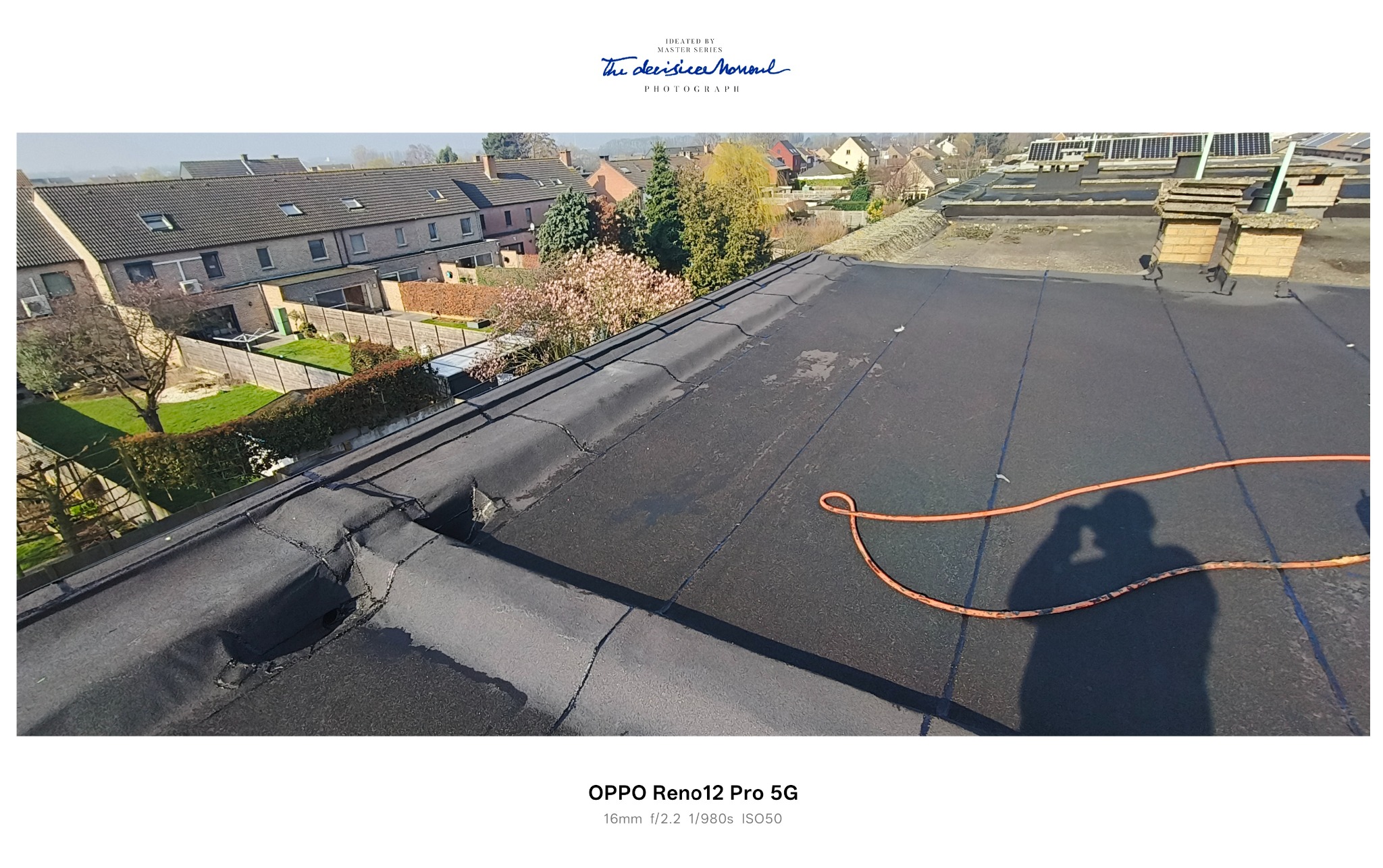 Płaski dach pokryty papą z widocznymi pęknięciami i uszkodzeniami. W tle zabudowa mieszkalna i drzewa. Na dachu leży pomarańczowy wąż i widać cień osoby.