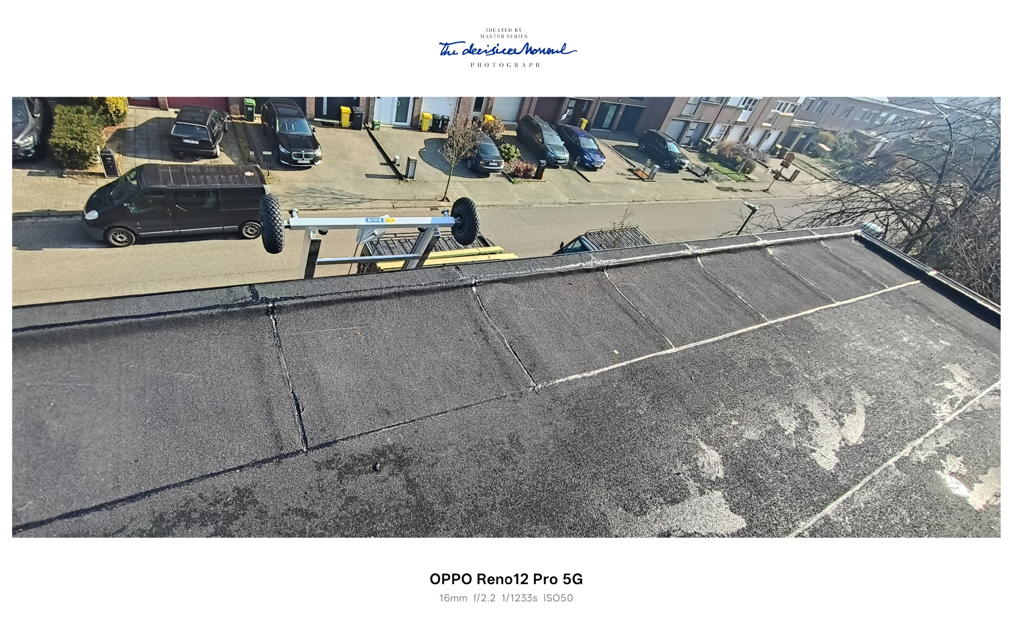 Widok z góry na płaski dach pokryty papą z widocznymi łączeniami, na tle ulicy z zaparkowanymi samochodami i budynkami mieszkalnymi. Widoczny podnośnik na dachu.