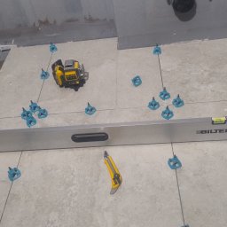 Przyrodzkibygg - Układanie płytek podłogowych z użyciem poziomicy, lasera krzyżowego i klipsów poziomujących. Widoczne narzędzia pracy i fragment dłoni pracownika.