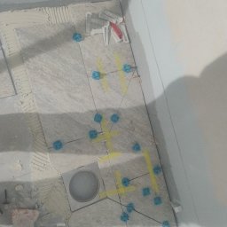Przyrodzkibygg - Układanie płytek podłogowych w łazience z użyciem niebieskich klinów i żółtej taśmy. Widoczny odpływ liniowy i narzędzia glazurnicze.