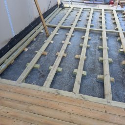 Przyrodzkibygg - Konstrukcja drewnianego tarasu w trakcie montażu: legary na podkładkach, częściowo ułożone deski tarasowe. Widoczne narzędzia i materiały budowlane.