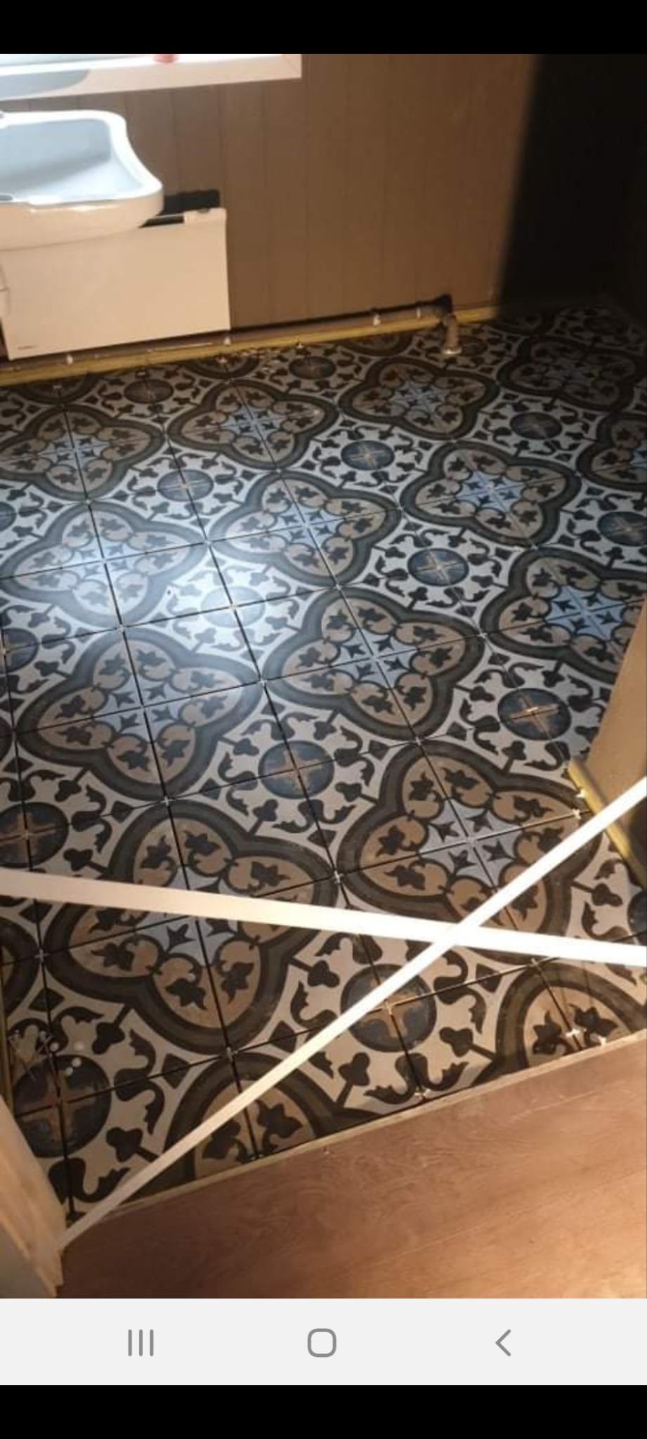 Ułożona podłoga z wzorzystych płytek w łazience, widoczna umywalka i fragment ściany. Dekoracyjny motyw płytek w odcieniach szarości i beżu.