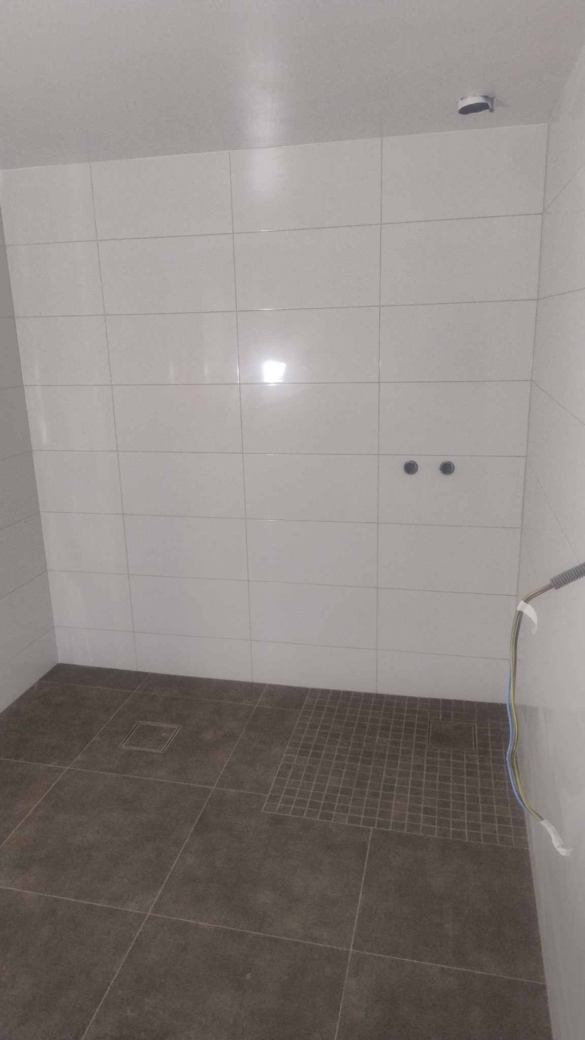 Wykończona kabina prysznicowa z brązowymi płytkami podłogowymi i białymi płytkami ściennymi. Widoczne odpływy liniowe i punktowe.