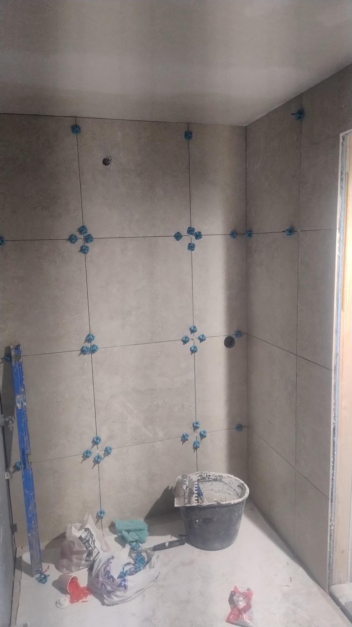 Ściana łazienki w trakcie wykańczania: szare płytki z niebieskimi krzyżykami dystansowymi, wiadro z zaprawą, drabina. Widoczne przewody i otwory instalacyjne.