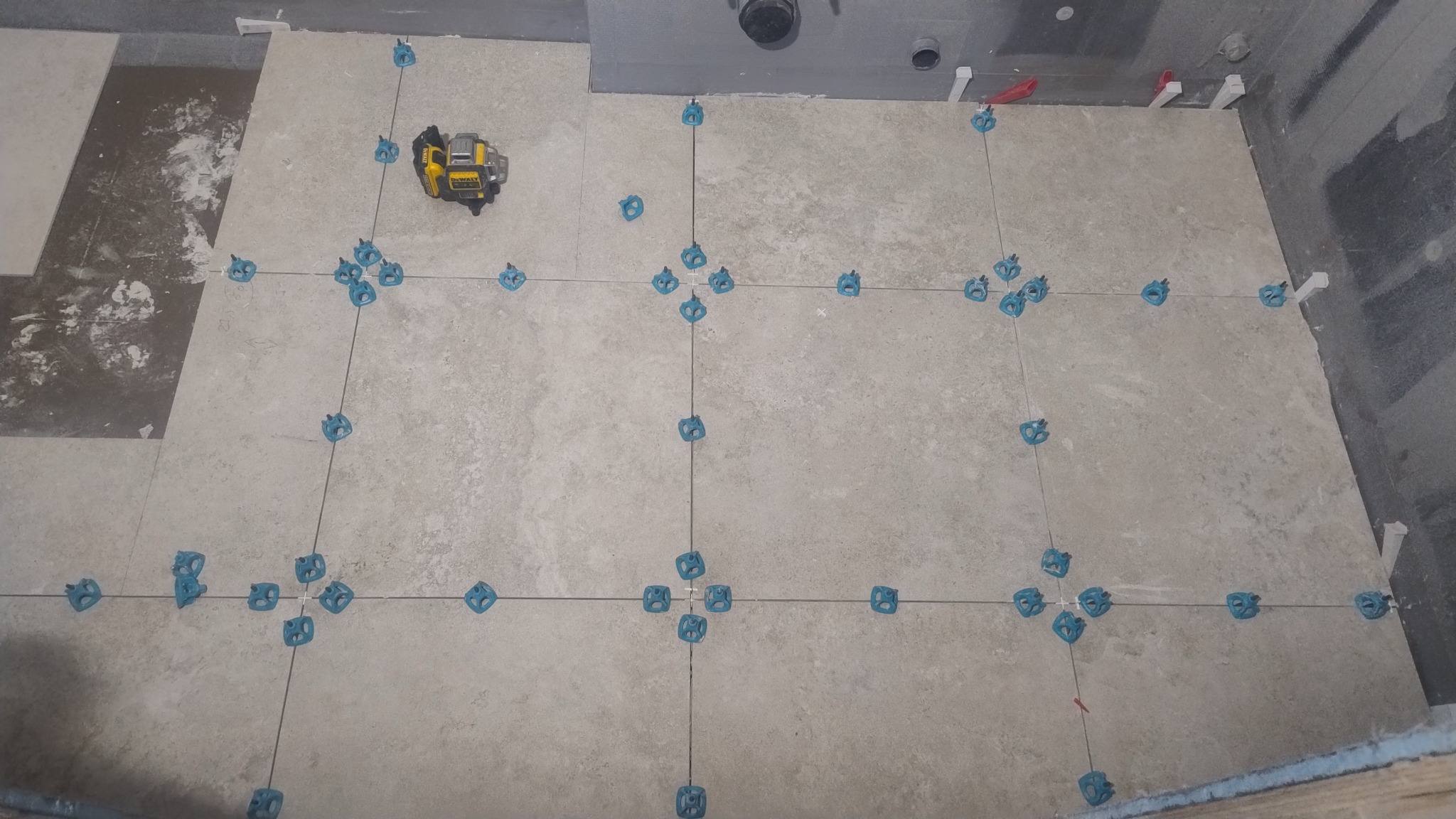 Układanie beżowych płytek podłogowych w łazience, z użyciem poziomicy laserowej i niebieskich klipsów do poziomowania. Widoczne fugi i fragmenty nieułożonych płytek.