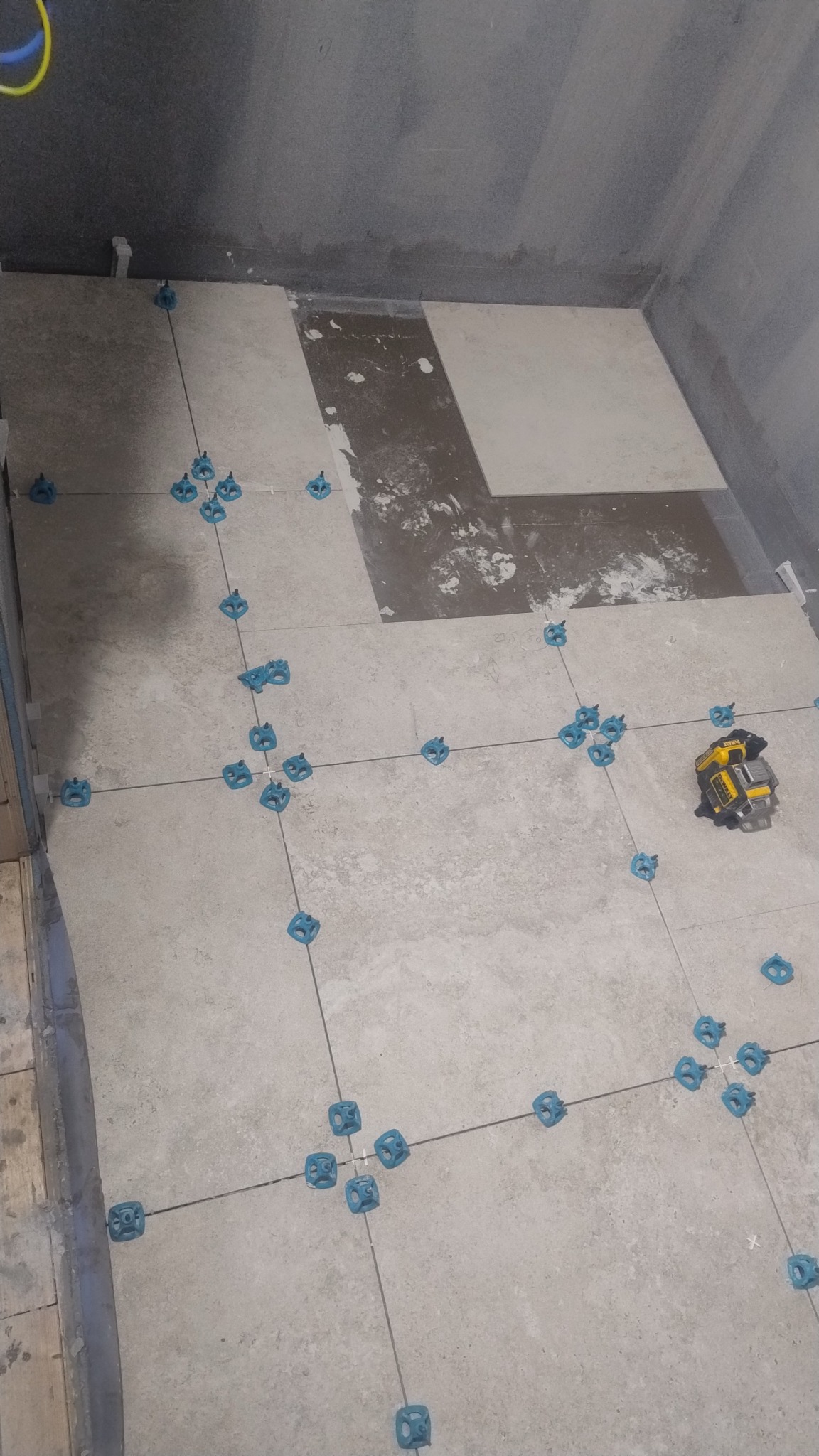 Układanie płytek podłogowych w łazience z użyciem poziomicy laserowej i systemu poziomowania płytek. Widok z góry na częściowo wyłożoną podłogę.