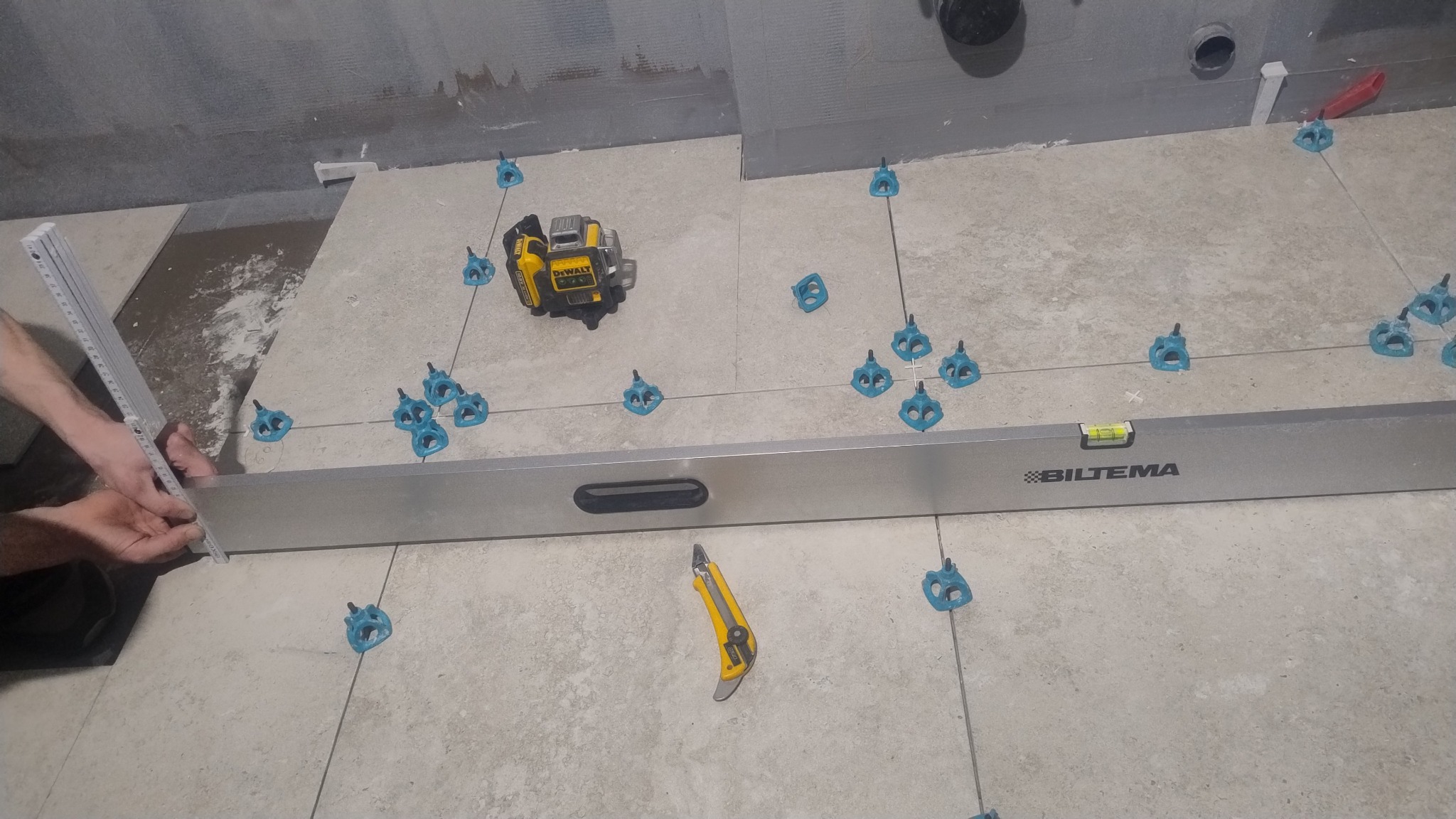 Układanie płytek podłogowych z użyciem poziomicy, lasera krzyżowego i klipsów poziomujących. Widoczne narzędzia pracy i fragment dłoni pracownika.