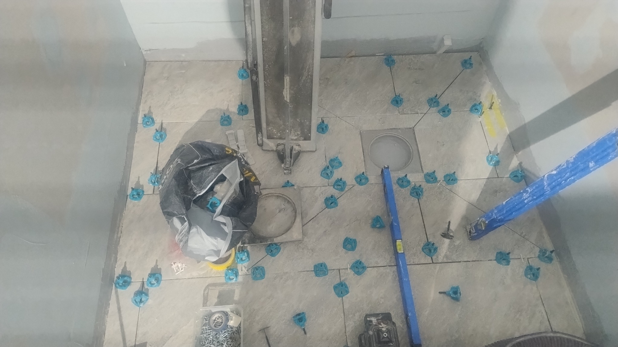 Układanie płytek podłogowych w łazience, widoczne narzędzia i poziomica. Etap prac z użyciem systemów poziomowania płytek. Szare płytki i niebieskie klipsy.