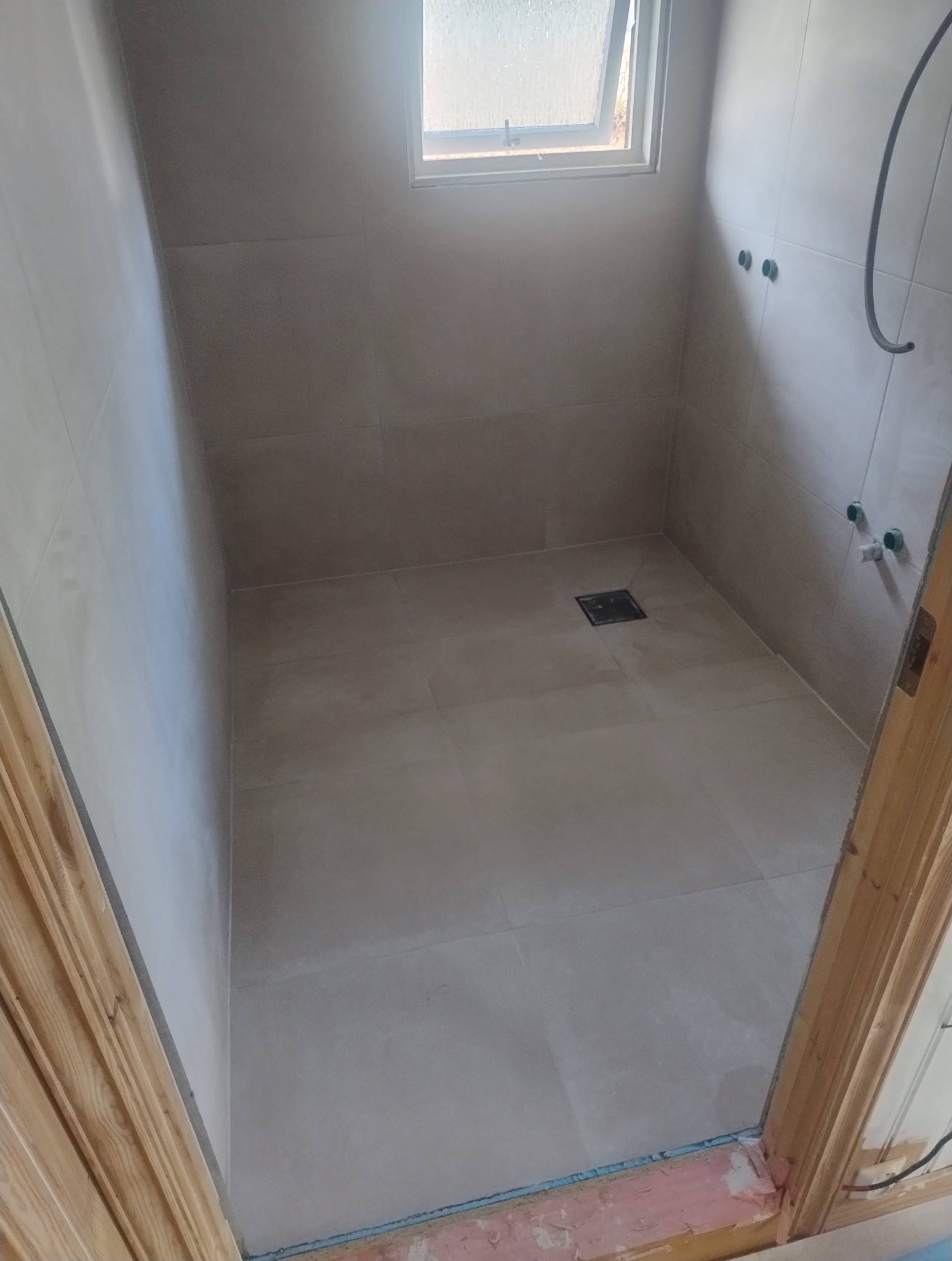 Wykończona kabina prysznicowa z beżowymi płytkami i odpływem liniowym. Małe okno doświetla wnętrze. Widoczne wystające rury instalacji wodnej.