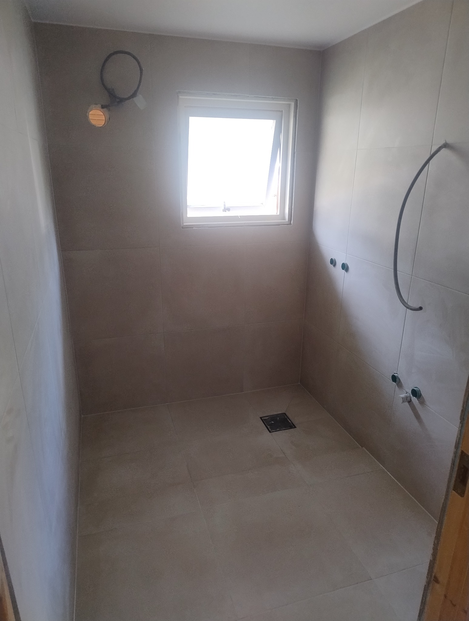 Wykończona łazienka z beżowymi płytkami, widoczne przyłącza wodne i okno. Minimalistyczny design z kratką odpływową w podłodze.