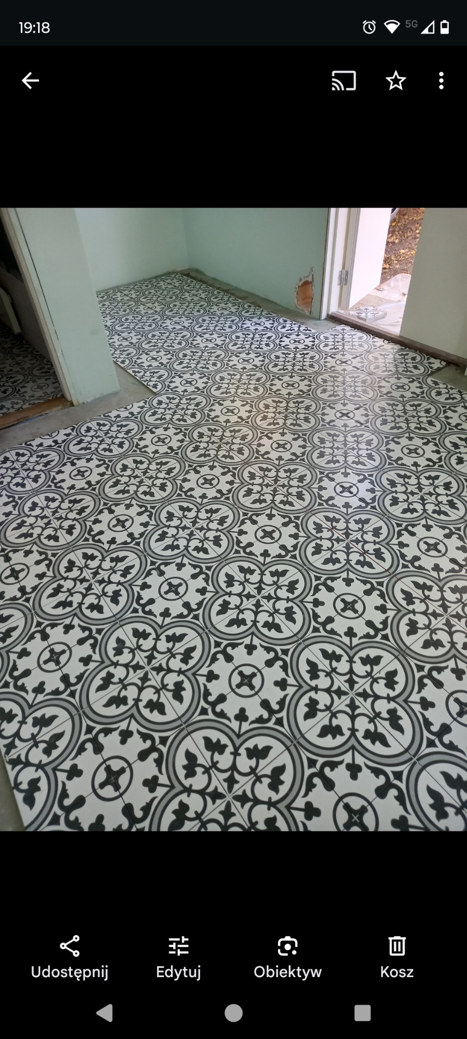 Wzorzysta, czarno-biała podłoga z płytek ceramicznych w pokoju. Widoczny fragment ściany w odcieniach bieli i otwarte drzwi z widokiem na zewnątrz.