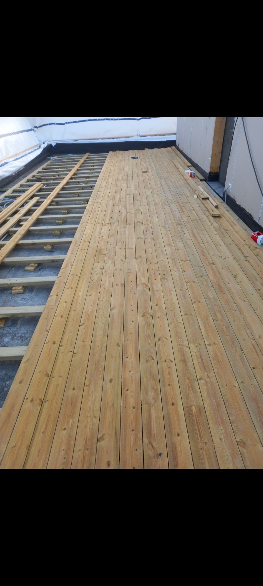 Montaż drewnianej podłogi tarasu: częściowo ułożone deski na legarach, widoczne materiały izolacyjne i elementy konstrukcyjne. Jasne drewno, etap budowy.