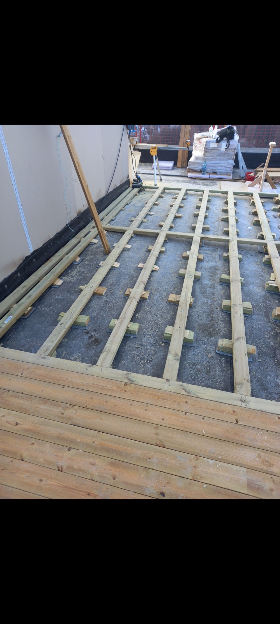Konstrukcja drewnianego tarasu w trakcie montażu: legary na podkładkach, częściowo ułożone deski tarasowe. Widoczne narzędzia i materiały budowlane.