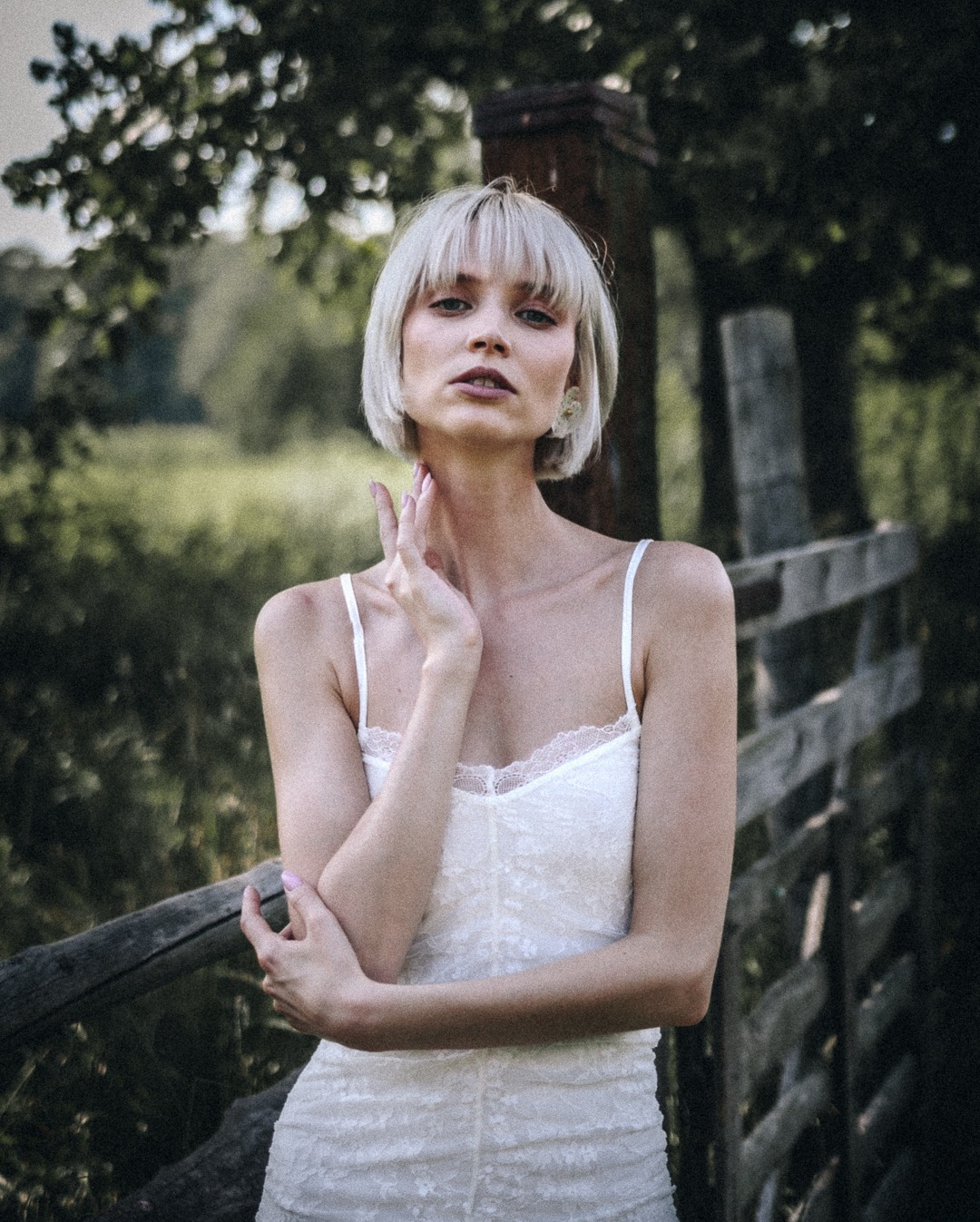 Portret kobiety w białej sukience na tle zieleni i drewnianego płotu. Delikatne światło, naturalna poza, subtelny makijaż. Ujęcie plenerowe, artystyczna fotografia.