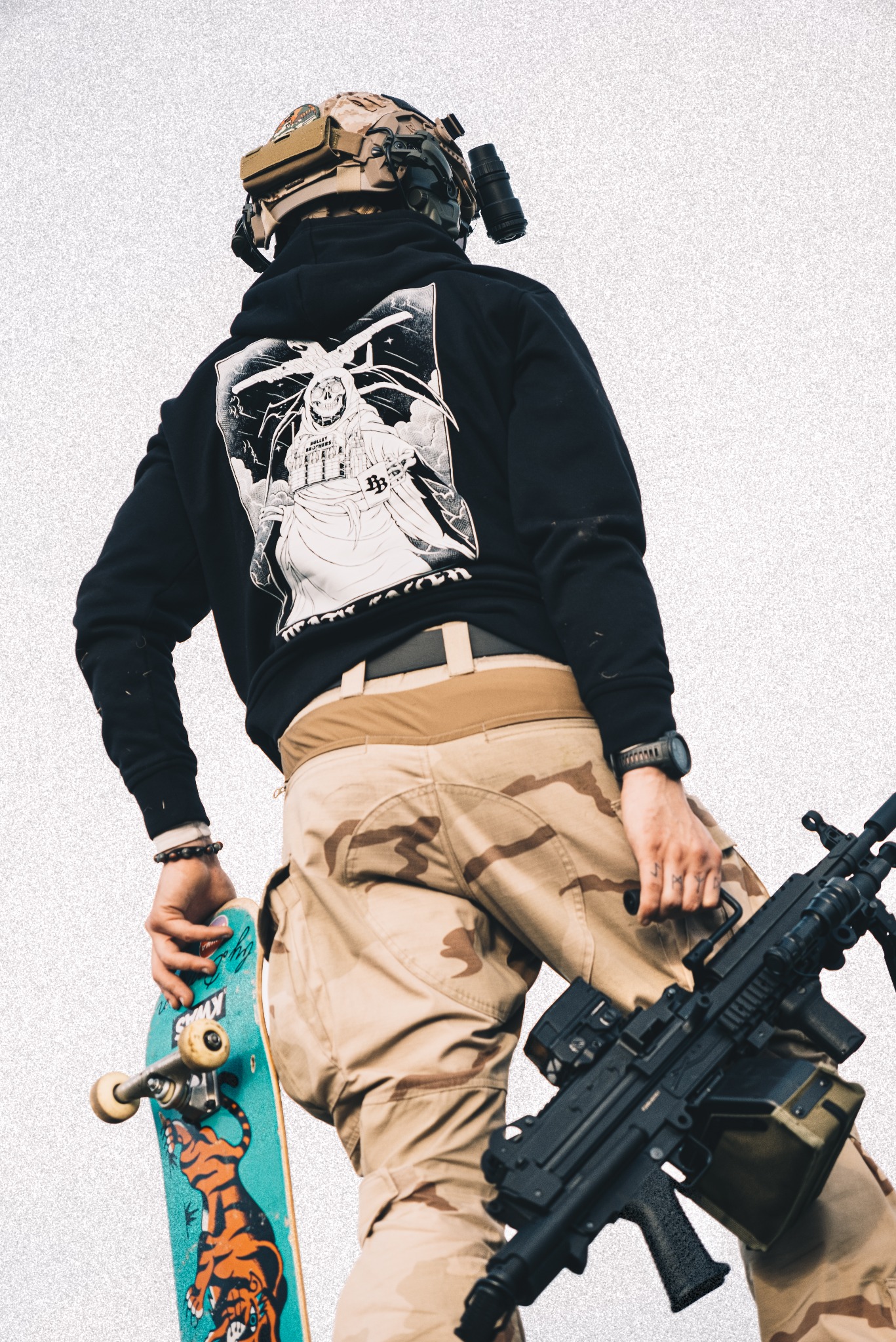 Osoba z tyłu w hełmie, bluzie z grafiką i spodniach moro trzyma deskorolkę i karabin maszynowy na tle jasnego tła. Nietypowe połączenie stylów.