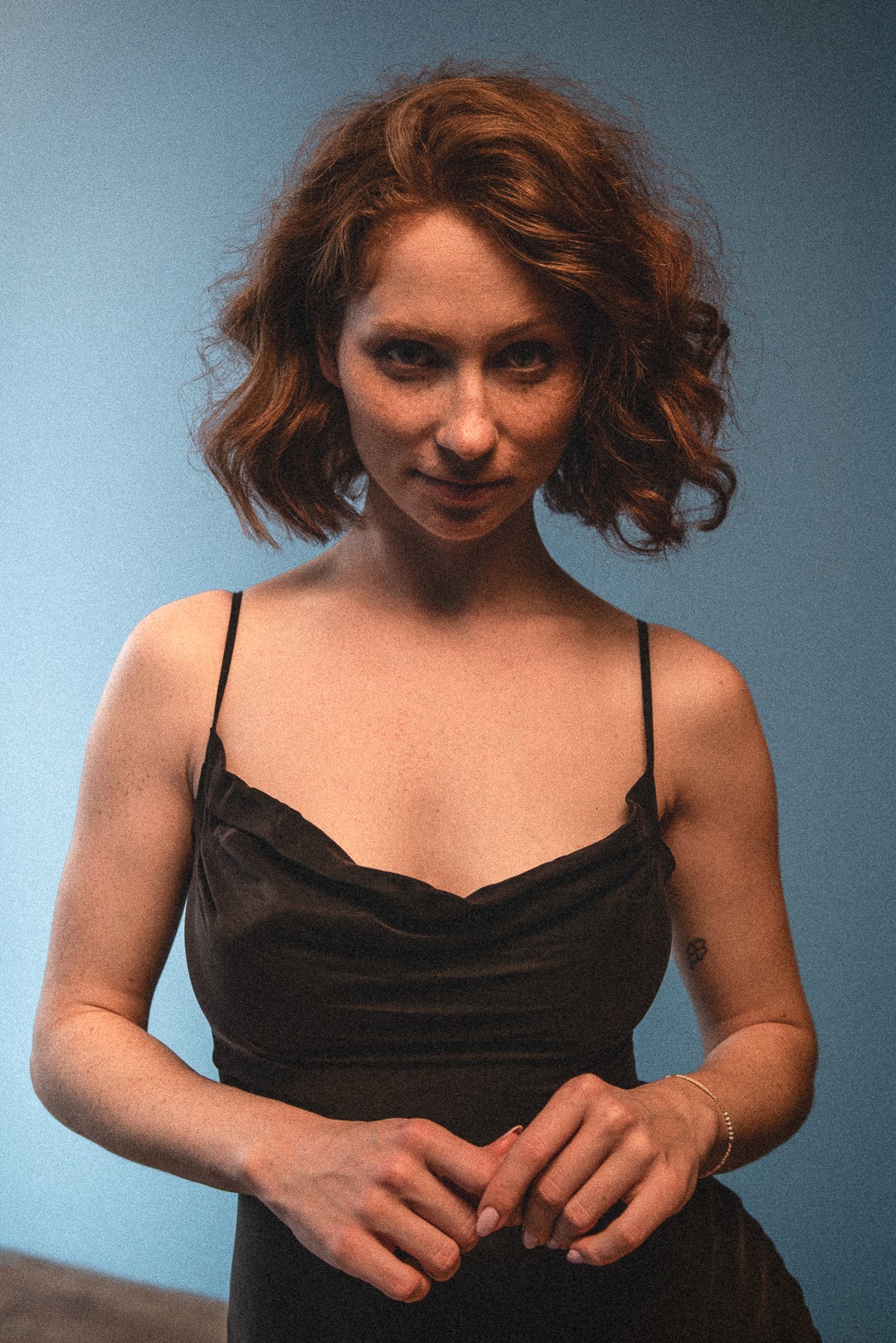 Portret kobiety o rudych włosach, w czarnej sukience na tle błękitnego tła. Delikatny uśmiech, widoczne piegi i subtelny tatuaż na ramieniu.