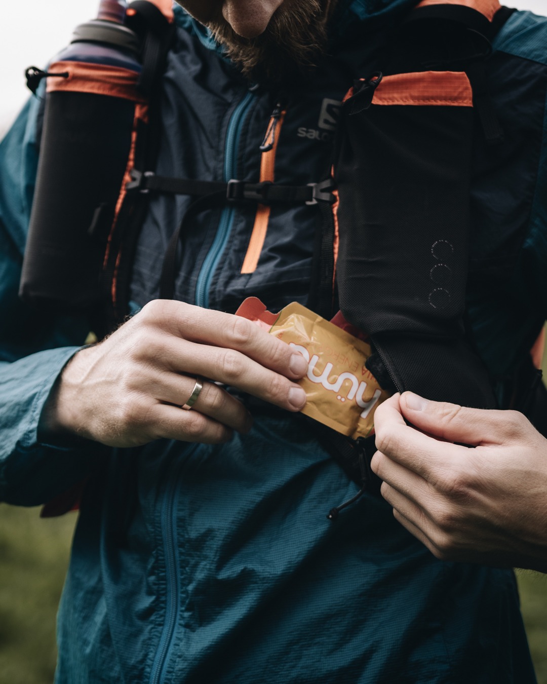 Mężczyzna w kurtce Salomon wyciąga żel energetyczny z kieszeni plecaka biegowego. Widoczne dłonie z obrączką, fragment brody i butelka z wodą.