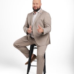 LIBERTY SPÓŁKA Z OGRANICZONĄ ODPOWIEDZIALNOŚCIĄ - Mężczyzna w beżowym garniturze siedzi na czarnym stołku, wskazując palcami w stronę widza. Białe tło, brązowe mokasyny, zegarek na ręce.