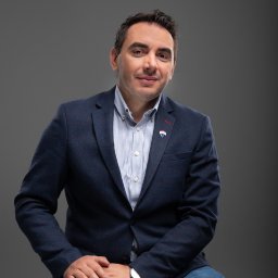 LIBERTY SPÓŁKA Z OGRANICZONĄ ODPOWIEDZIALNOŚCIĄ - Portret uśmiechniętego mężczyzny w marynarce z przypinką, siedzącego na krześle, na jednolitym szarym tle. Ubrany w jeansy i koszulę w paski, na ręku zegarek.