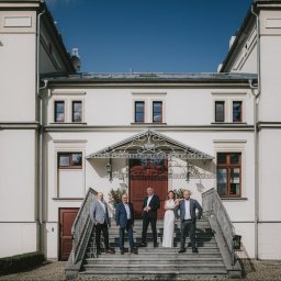 LIBERTY SPÓŁKA Z OGRANICZONĄ ODPOWIEDZIALNOŚCIĄ - Elewacja odnowionego pałacyku z grupą osób na schodach wejściowych. Jasna fasada, ciemne okna, błękitne niebo. Reprezentacyjna architektura po renowacji.