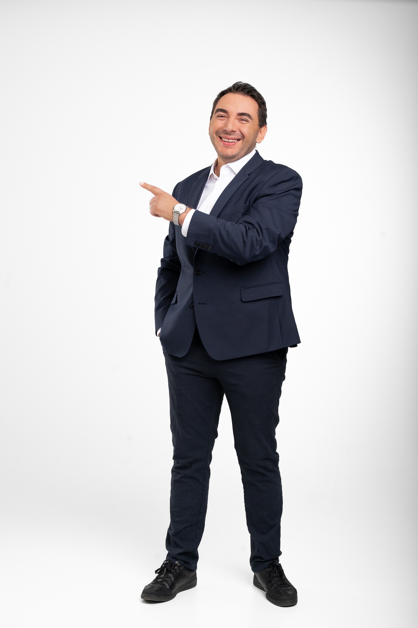 Uśmiechnięty mężczyzna w granatowym garniturze i białej koszuli, z zegarkiem, wskazuje palcem w lewo na białym tle. Wygląda profesjonalnie i pewnie.