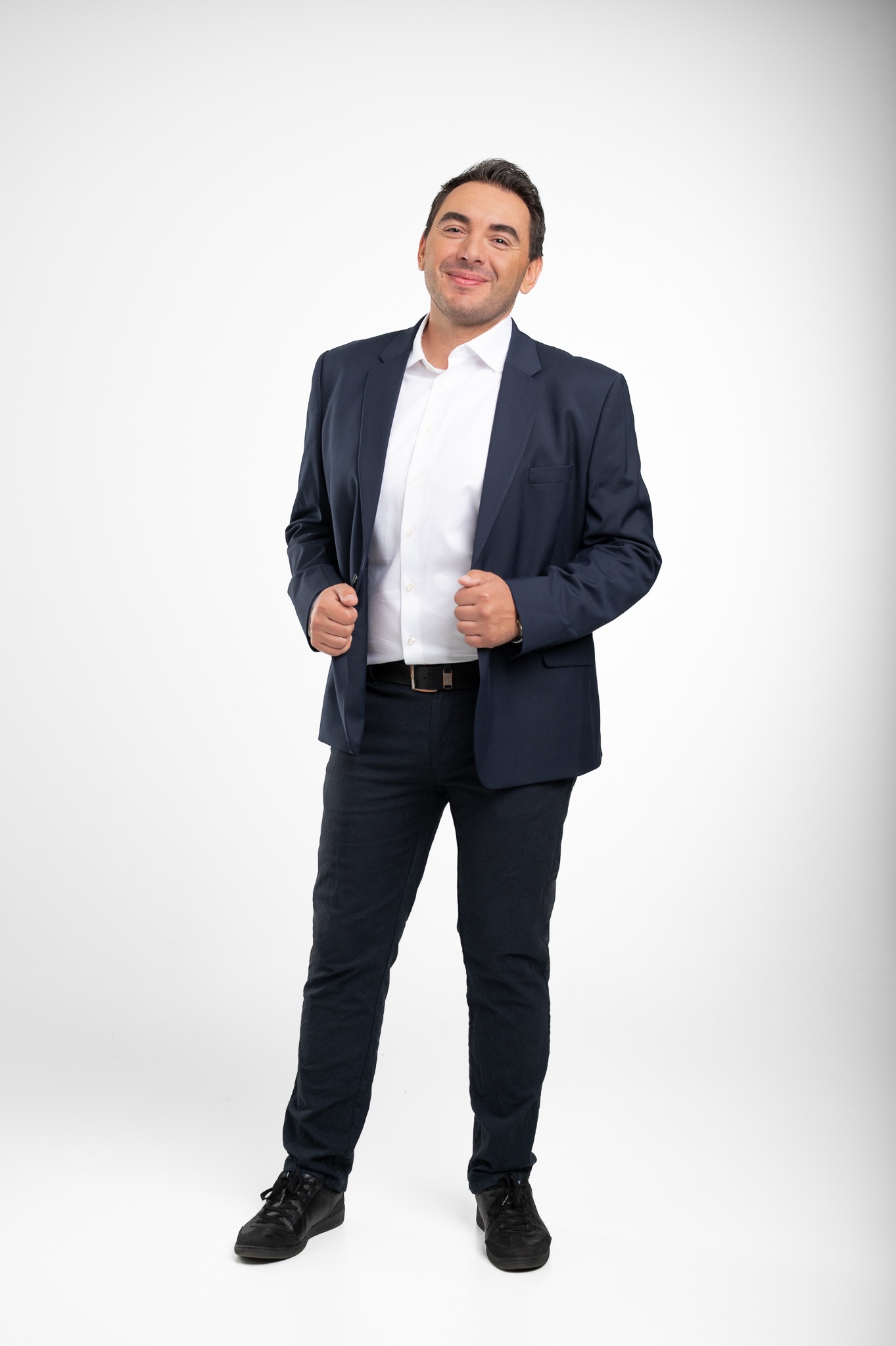 Uśmiechnięty mężczyzna w granatowym garniturze i białej koszuli, stojący na jasnym tle. Prezentacja wizerunku doradcy lub agenta.