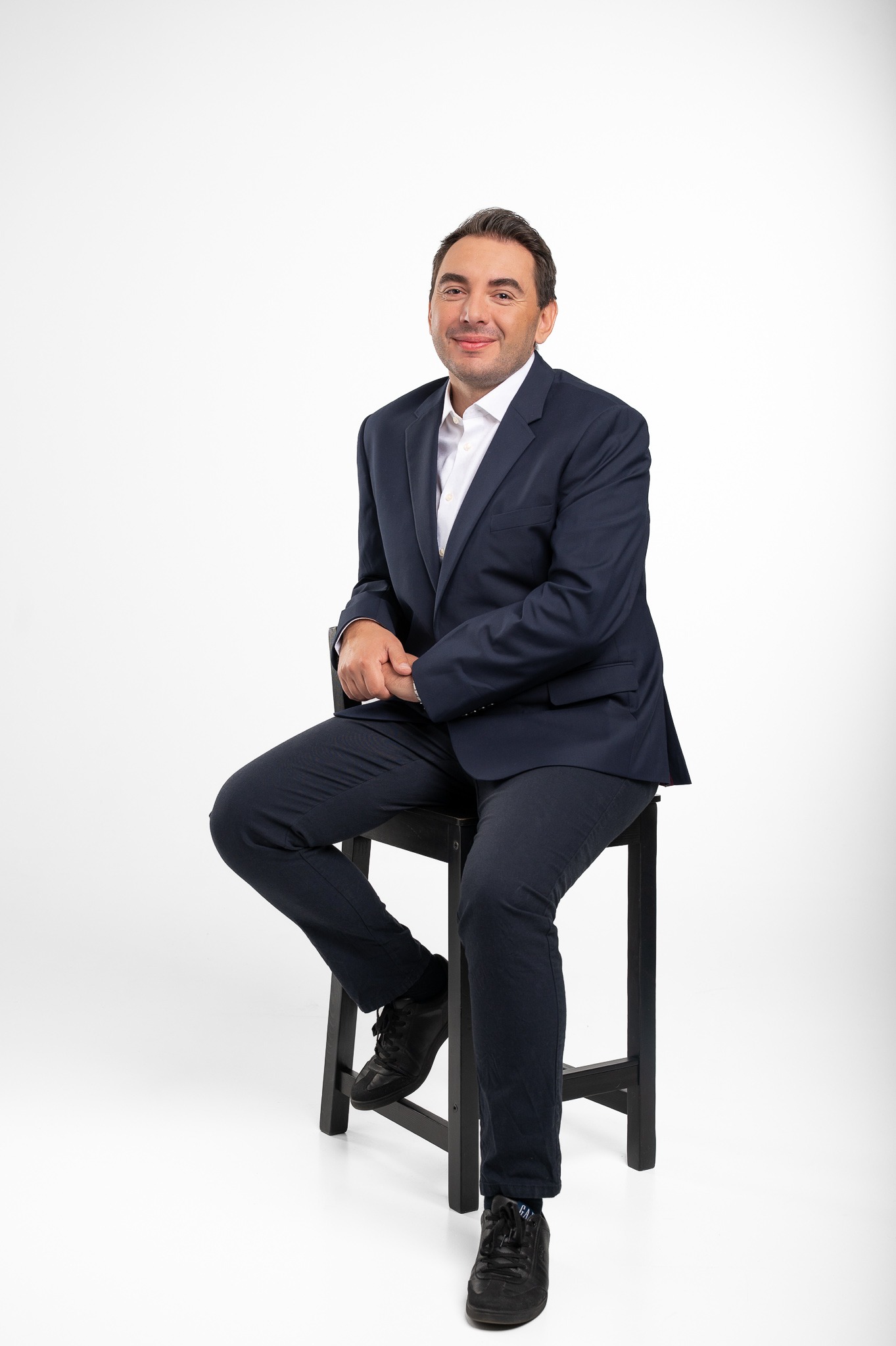 Mężczyzna w granatowym garniturze i białej koszuli siedzi na czarnym stołku na białym tle, spoglądając w obiektyw z lekkim uśmiechem.