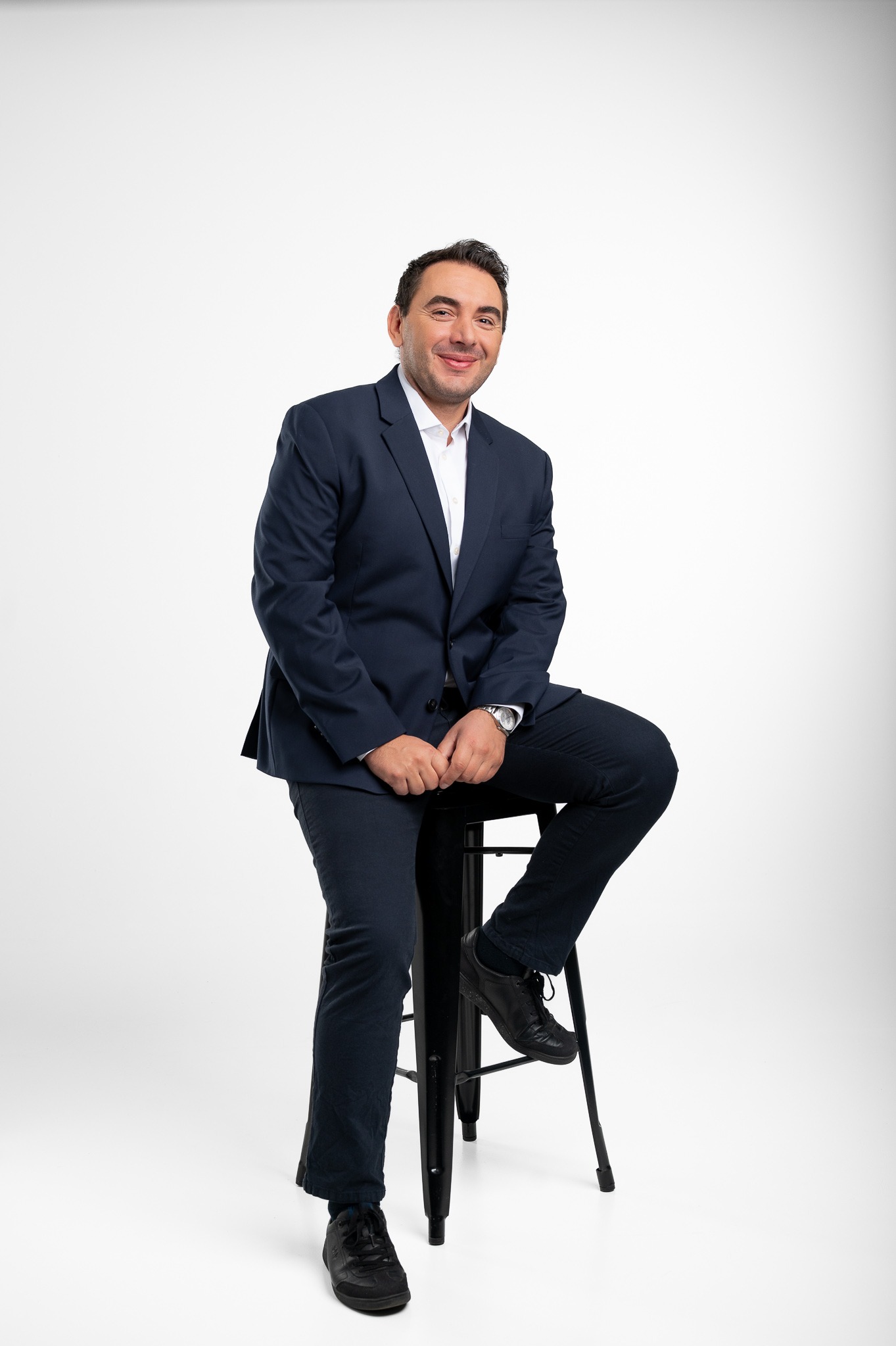 Portret uśmiechniętego mężczyzny w granatowym garniturze, siedzącego na czarnym stołku barowym na jasnym tle. Widoczny zegarek na ręce.