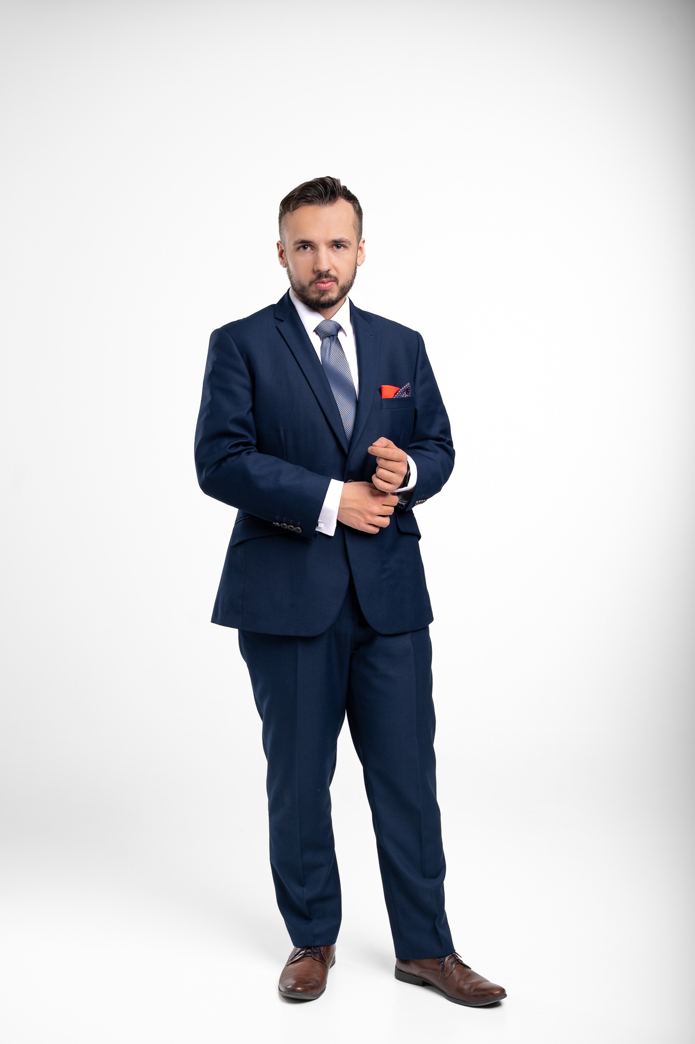 Mężczyzna w granatowym garniturze na białym tle, zapinający mankiet koszuli. Widoczna poszetka z wzorem. Elegancki wygląd, profesjonalny wizerunek.