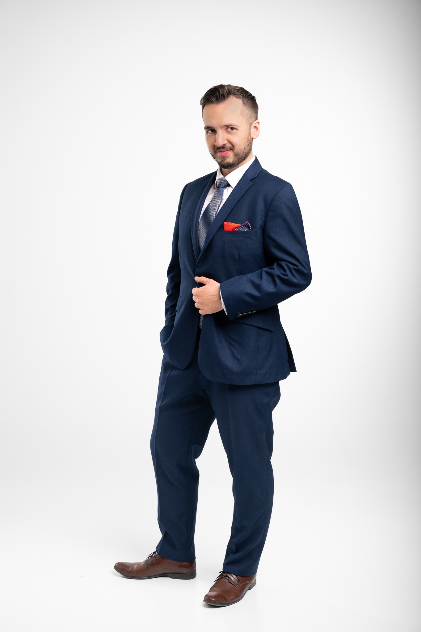 Mężczyzna w granatowym garniturze, z czerwoną poszetką i brązowymi butami, stoi na jasnym tle. Uśmiecha się, trzymając rękę na klapie marynarki. Elegancki wygląd.