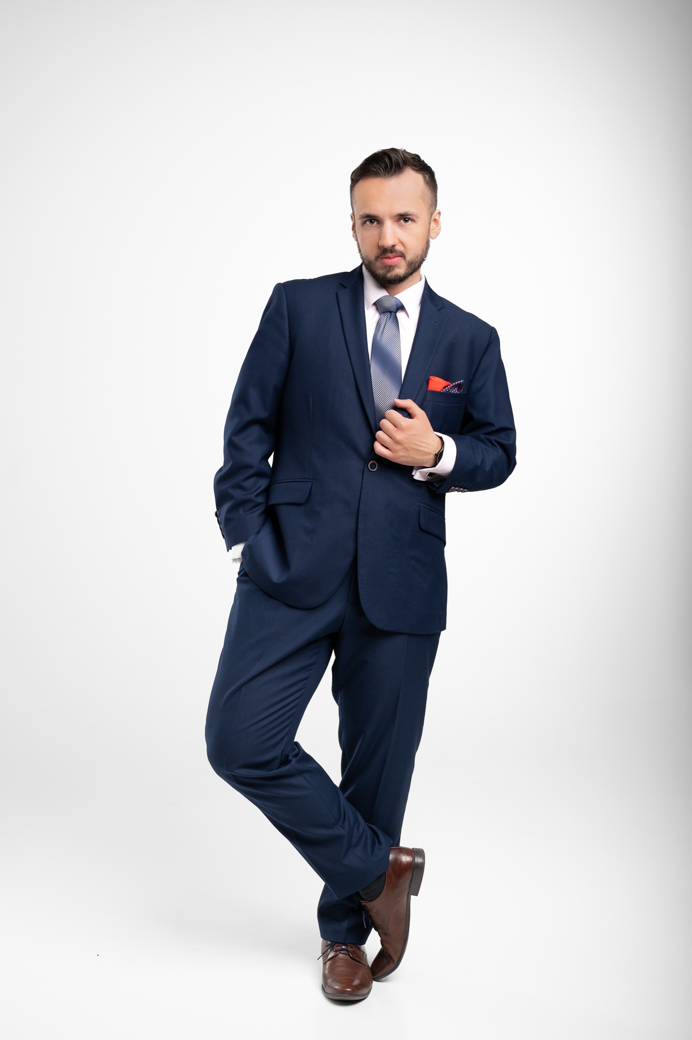 Mężczyzna w granatowym garniturze, błękitnym krawacie i brązowych butach, pozuje na białym tle, trzymając dłoń na klapie marynarki. W kieszonce widoczna poszetka.