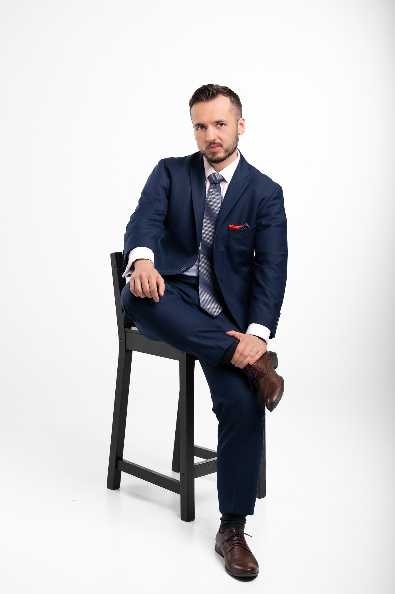 Mężczyzna w granatowym garniturze, szarym krawacie i brązowych butach siedzi na czarnym krześle na białym tle, prezentując profesjonalny wizerunek.