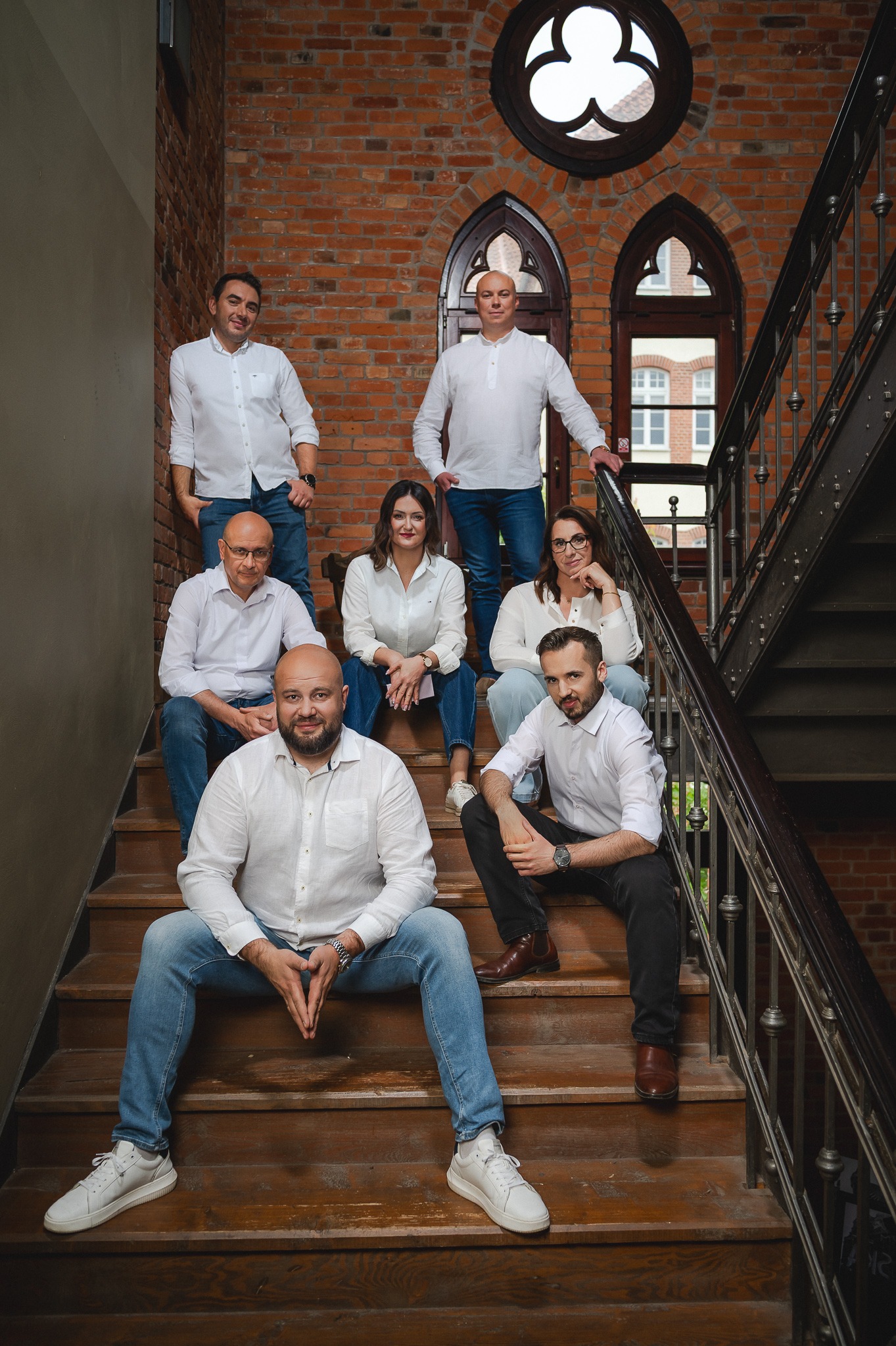 Zespół finansowy pozuje na schodach z ceglanym tłem i gotyckimi oknami. Wszyscy ubrani w białe koszule i jeansy, tworząc spójny, profesjonalny wizerunek.