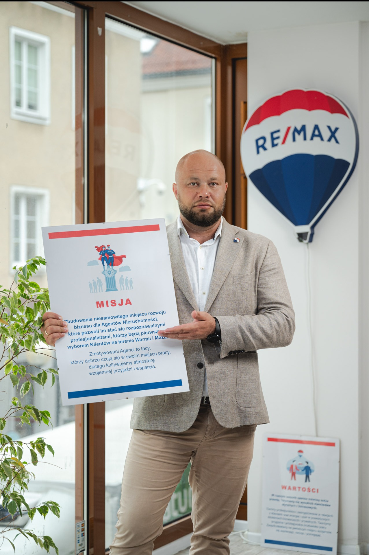 Agent nieruchomości prezentuje misję firmy na tle biura z logo RE/MAX i rośliną doniczkową. Mężczyzna w beżowej marynarce i spodniach.