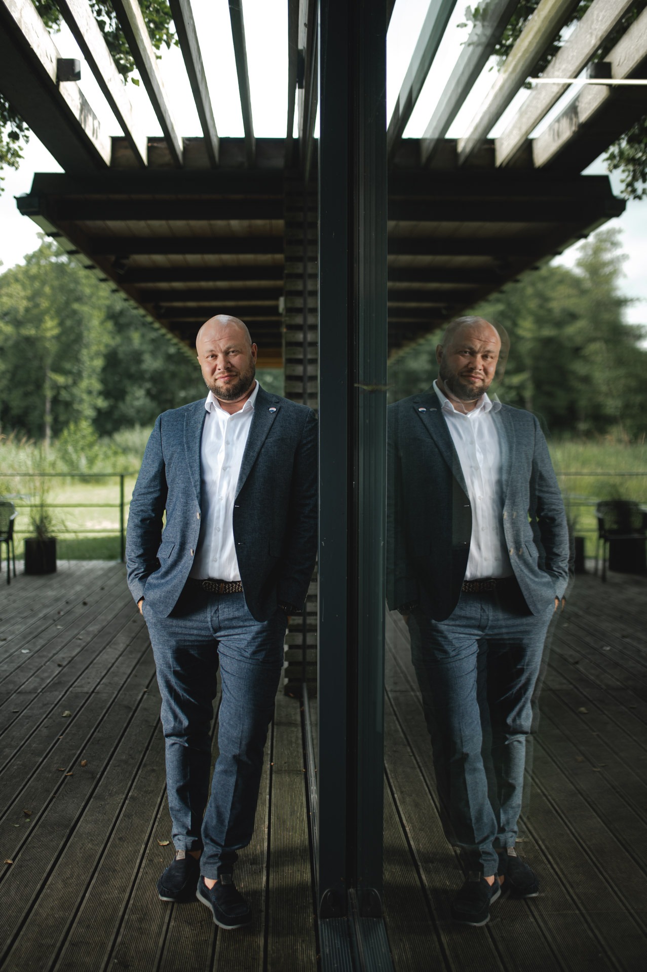 Mężczyzna w garniturze, oparty o szklaną ścianę z odbiciem, na drewnianym tarasie pod zadaszeniem. Ujęcie z dołu podkreśla sylwetkę i otoczenie.