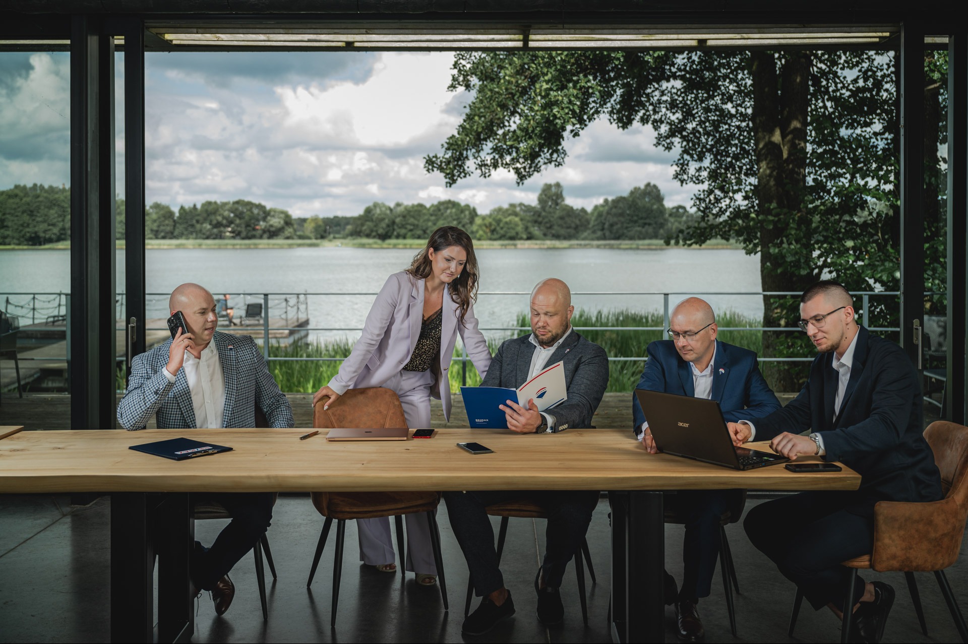 Grupa osób w garniturach siedzi przy stole z laptopami i dokumentami na tle jeziora. Jedna osoba rozmawia przez telefon, inna czyta dokument.