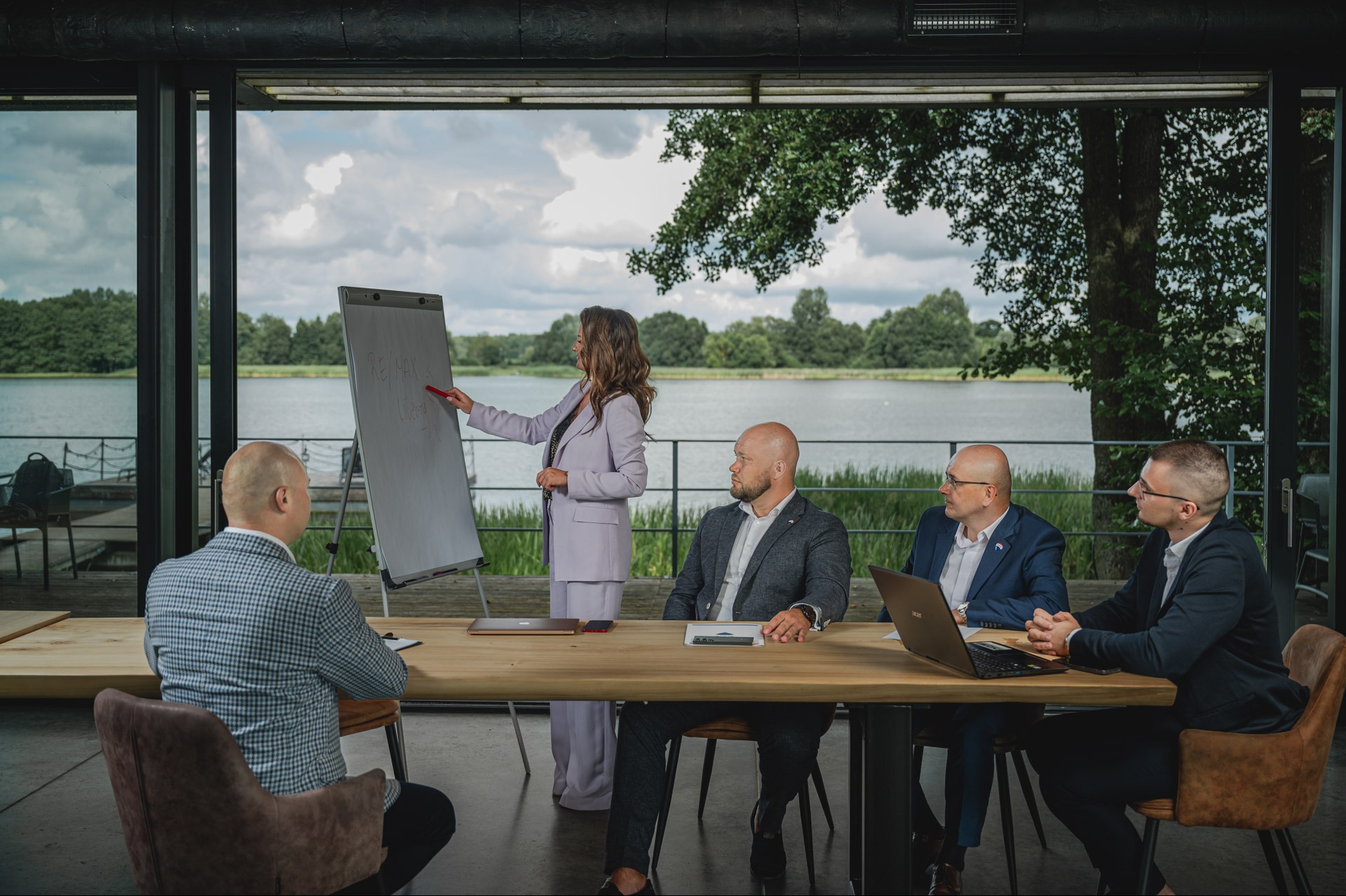 Prezentacja biznesowa nad jeziorem: kobieta w garniturze omawia dane na flipcharcie, słuchają jej mężczyźni w garniturach, widok na jezioro w tle.
