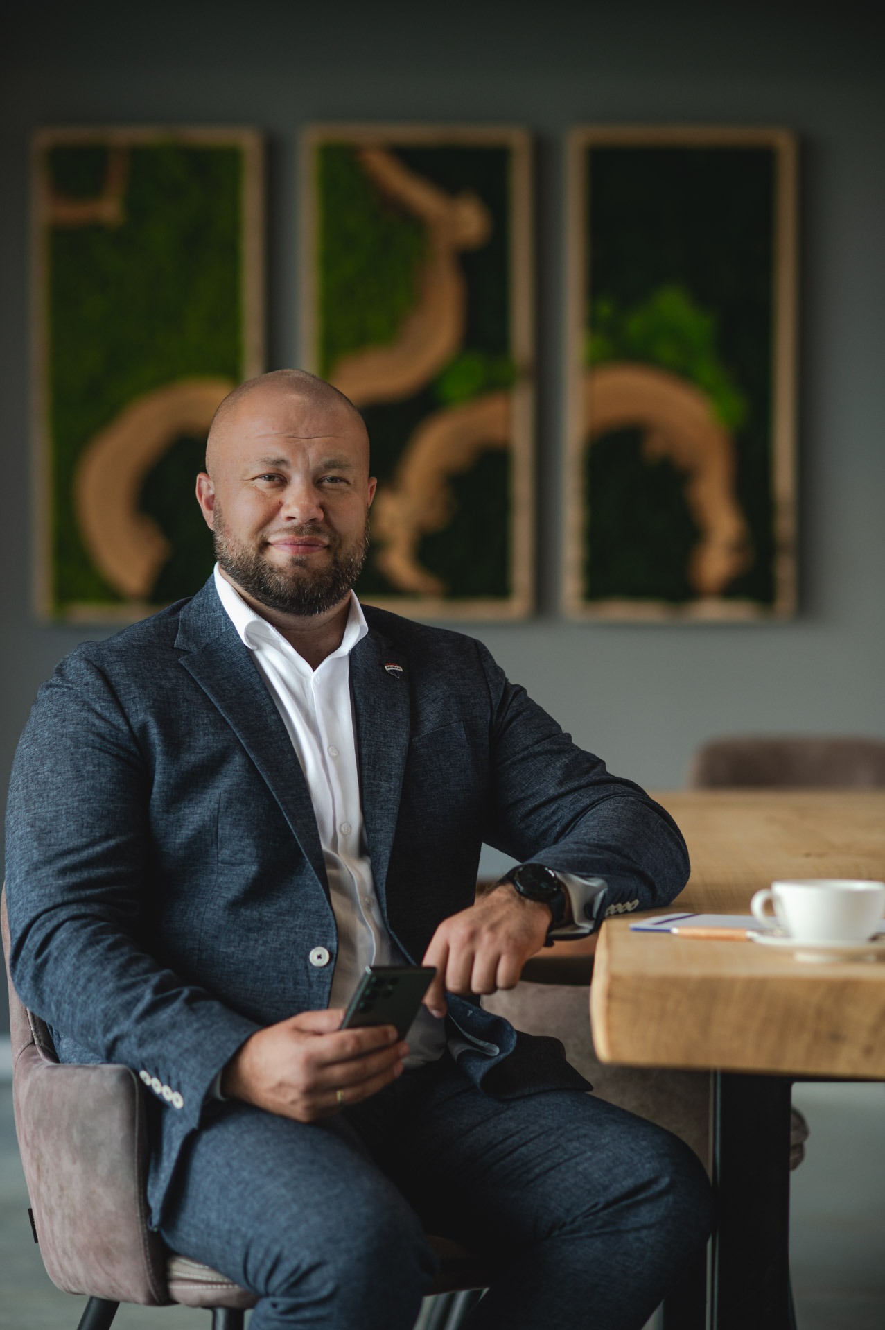 Mężczyzna w garniturze siedzi przy stole z telefonem, w tle dekoracje z mchu. Reprezentant agencji nieruchomości lub doradca finansowy.