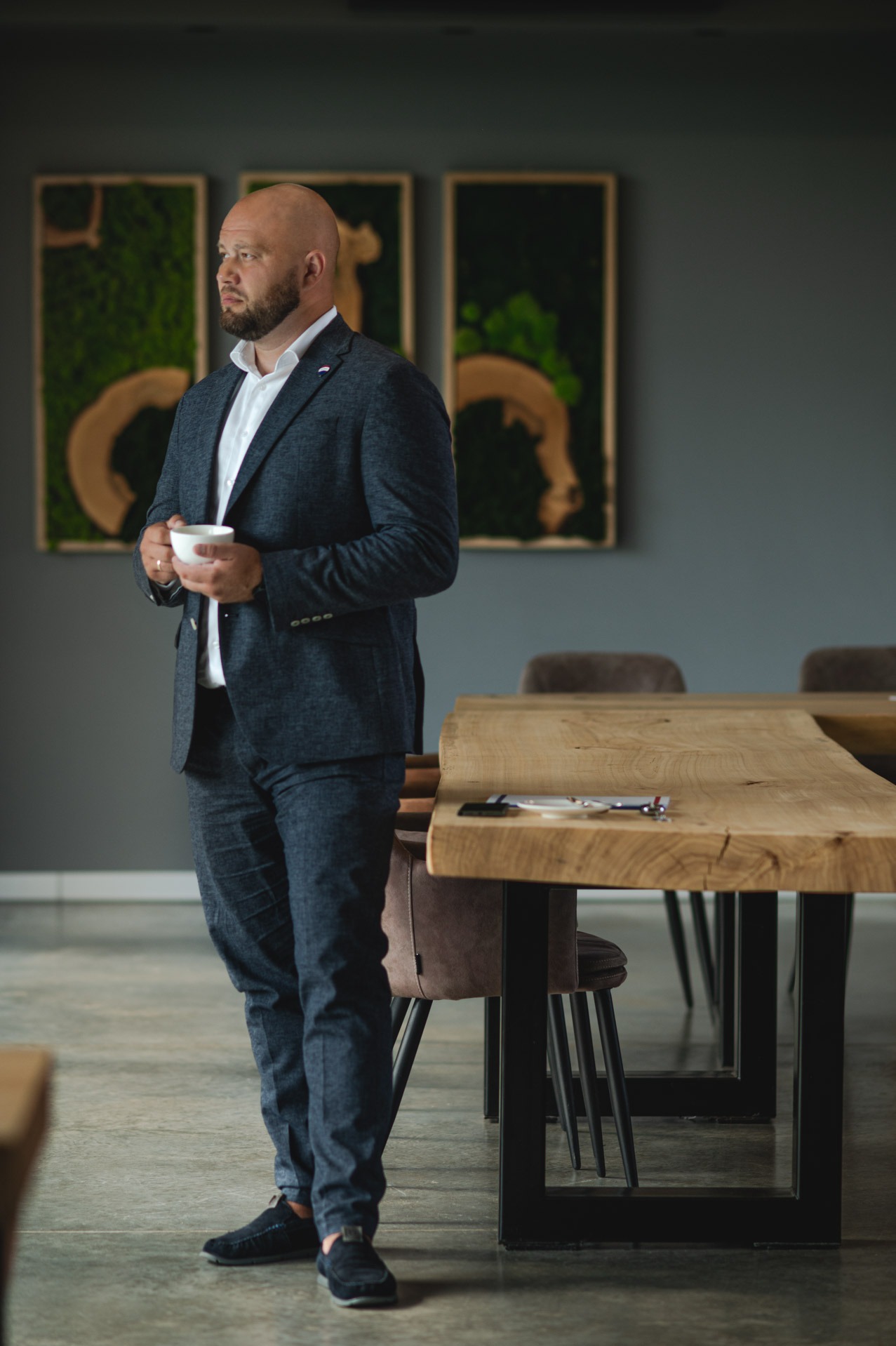 Mężczyzna w garniturze pije kawę w nowoczesnym biurze z drewnianym stołem i zielonymi obrazami na ścianie. Spokojna atmosfera, biznesowy styl.