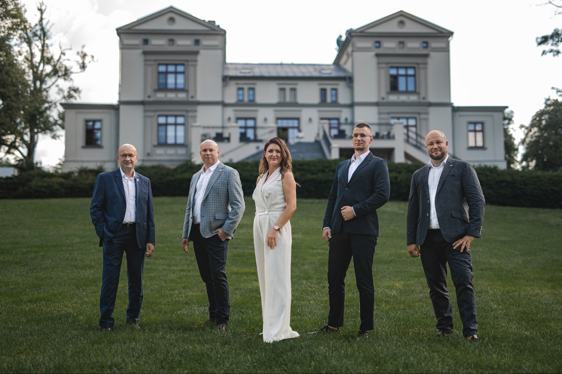 Zespół agentów nieruchomości pozuje na tle eleganckiego budynku. Kobieta w białym kombinezonie wyróżnia się na tle mężczyzn w garniturach. Ujęcie z szerokim kadrem.