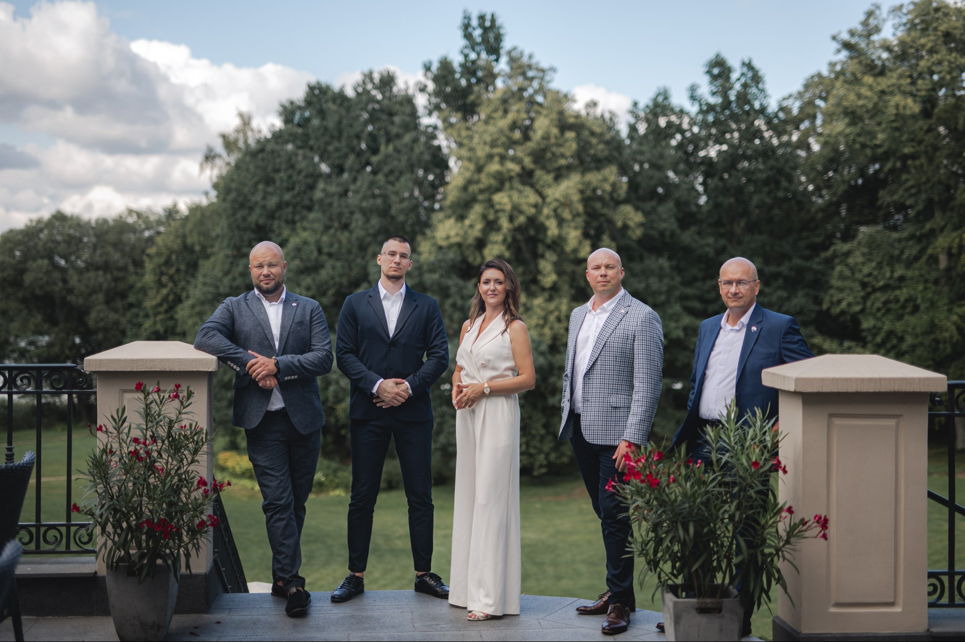 Grupa pięciu elegancko ubranych osób (czterech mężczyzn i kobieta) pozuje na zewnątrz, na tle zieleni i balustrady z kwiatami w donicach.