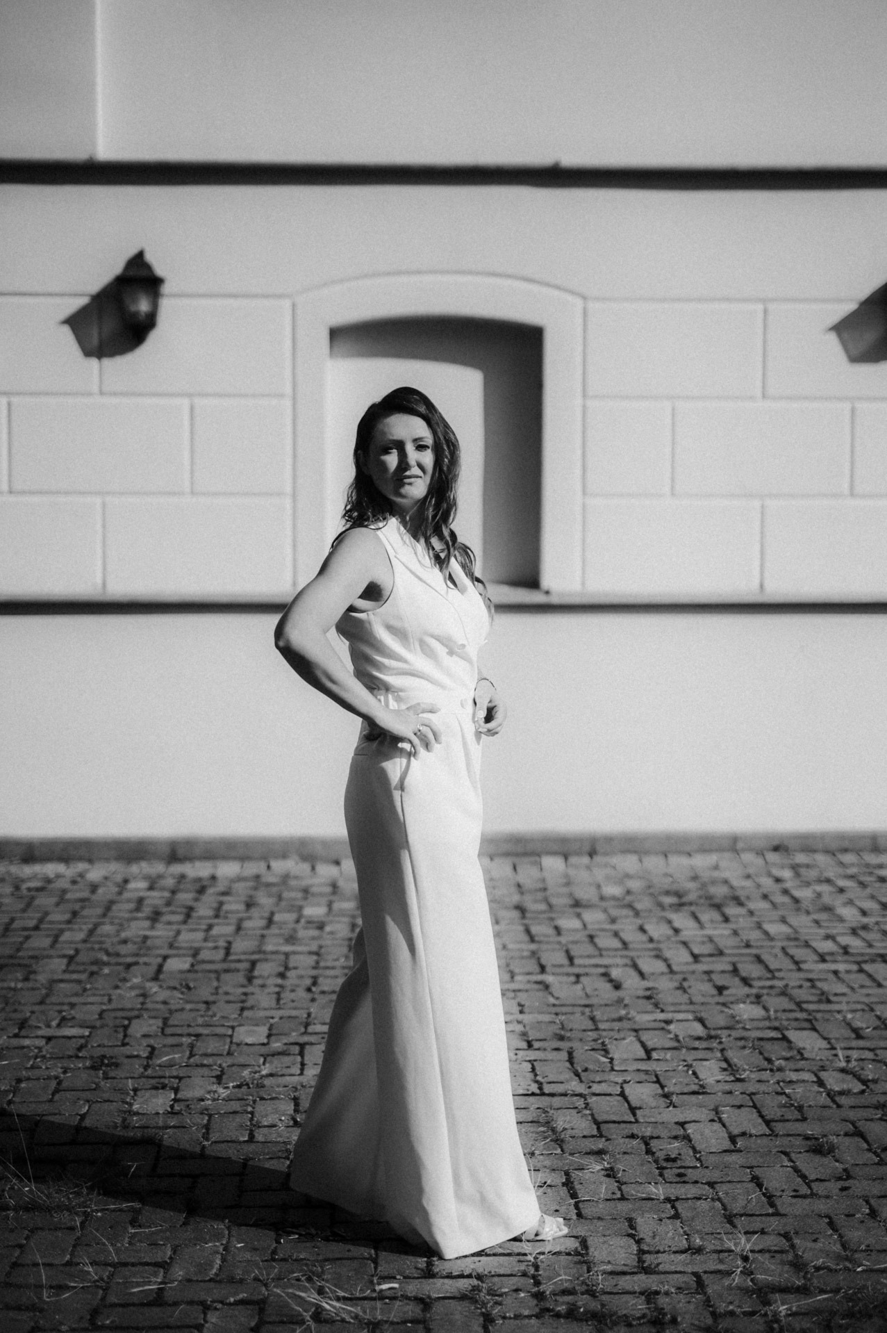 Czarno-białe zdjęcie kobiety w białym kombinezonie stojącej na brukowanej powierzchni przed budynkiem z lampami i małym oknem.