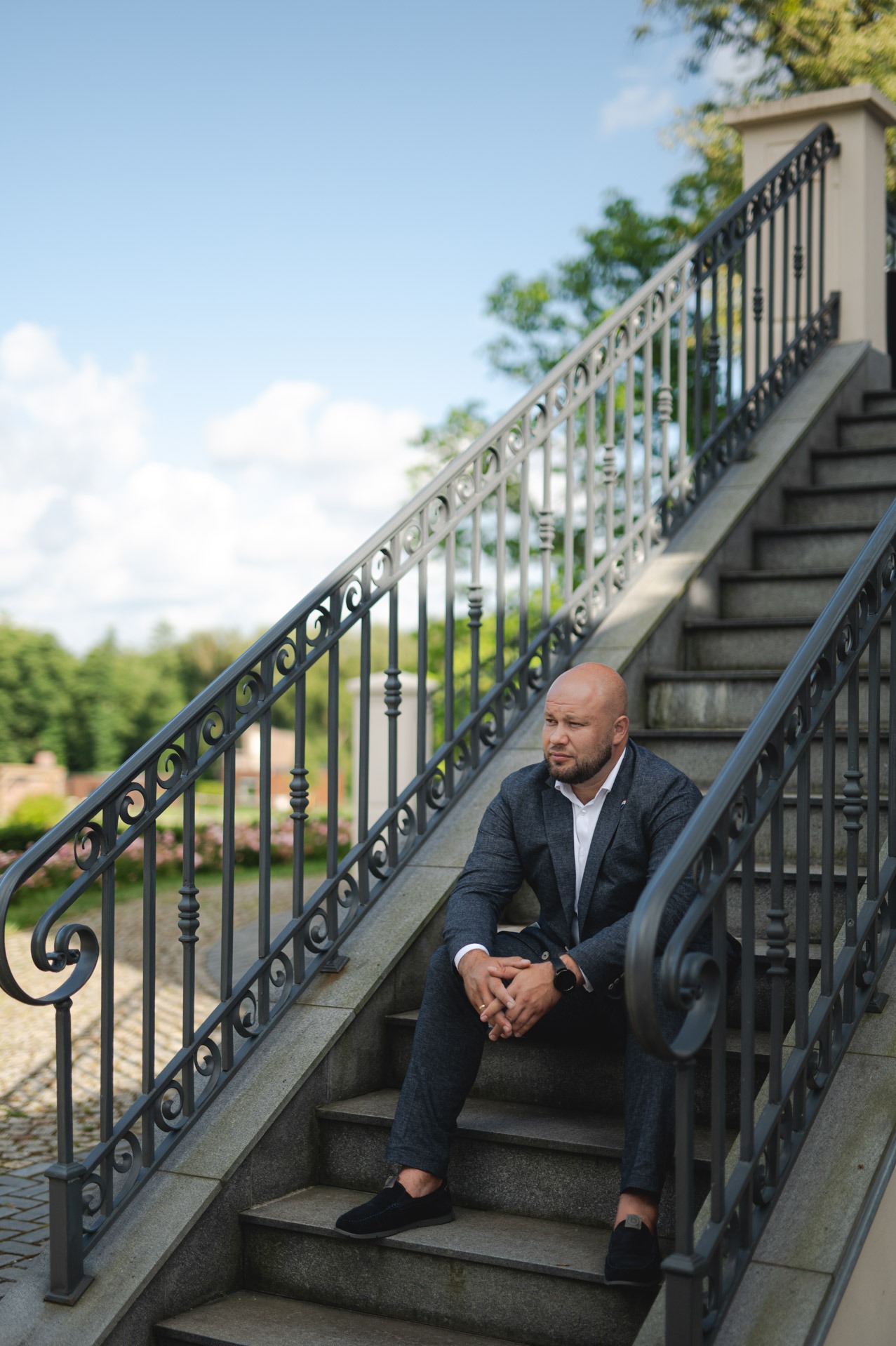 Mężczyzna w garniturze siedzi na kamiennych schodach z ozdobną, metalową balustradą, spoglądając w bok. W tle zieleń drzew i błękitne niebo.