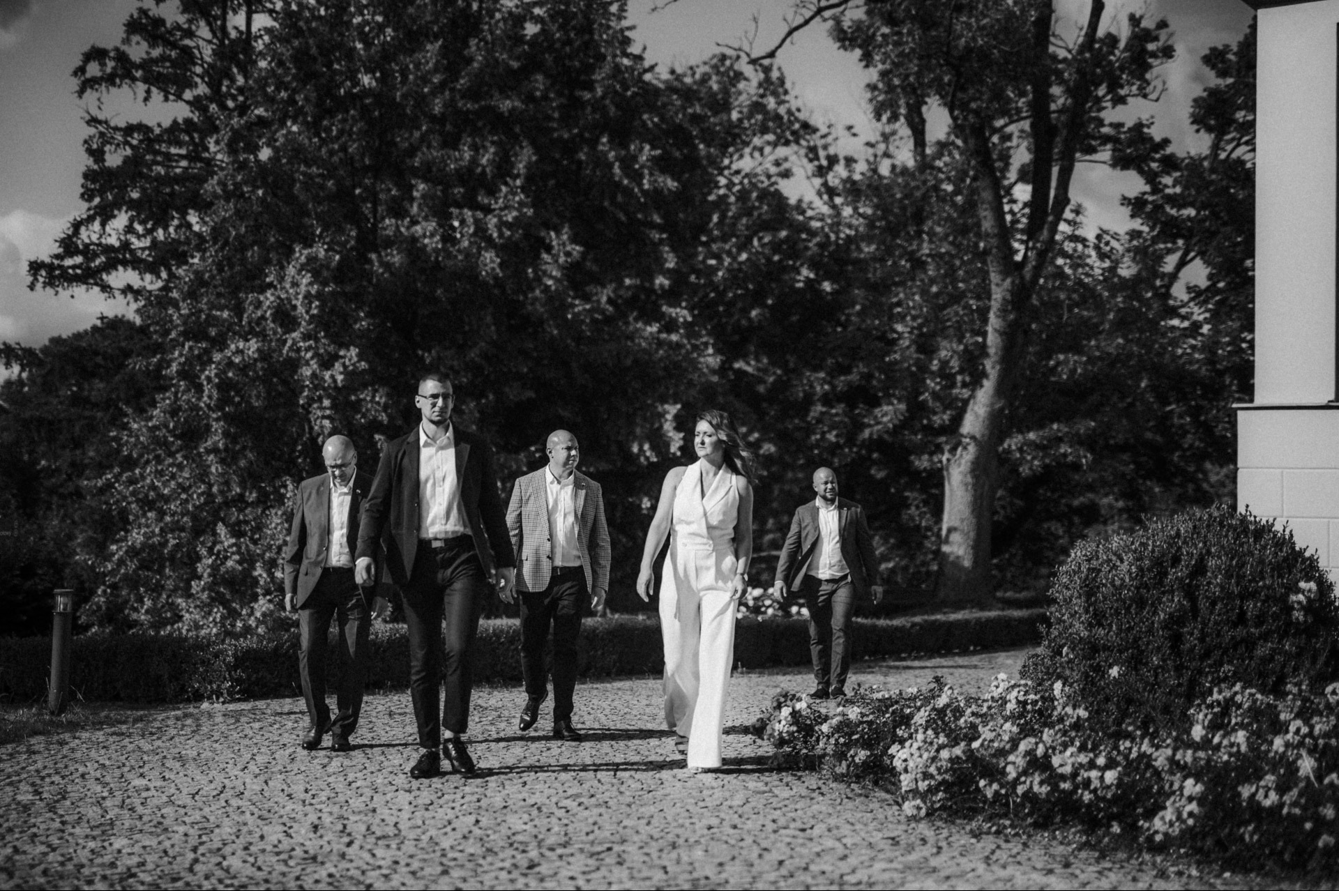Czarno-białe zdjęcie grupy osób (kobieta w białym kombinezonie i mężczyźni w garniturach) idących po brukowanej ścieżce w parku.