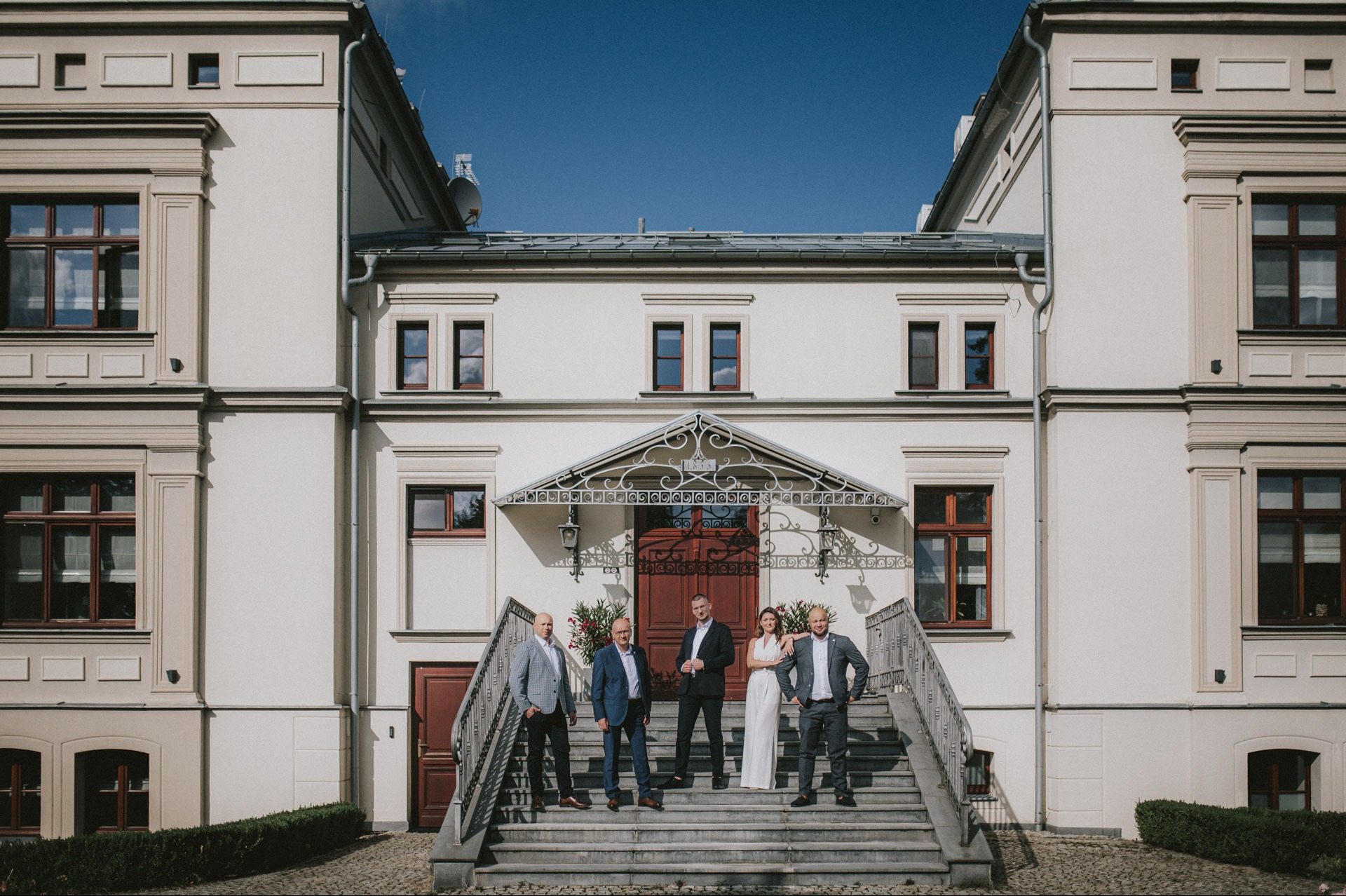 Elewacja odnowionego pałacyku z grupą osób na schodach wejściowych. Jasna fasada, ciemne okna, błękitne niebo. Reprezentacyjna architektura po renowacji.