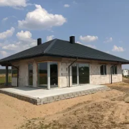 Niedokończony dom z białego pustaka z ciemnym dachem na tle błękitnego nieba z chmurami, widoczny z zewnątrz, z fragmentem tarasu i dwoma kominami.