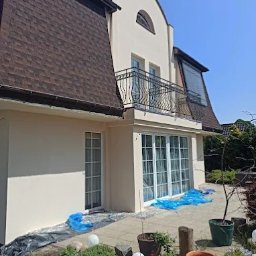 F.H.U Malors Marcin Słomski - Elewacja domu po tynkowaniu maszynowym, widok z ogrodu. Zabezpieczone folią okna tarasowe i roślinność. Brązowy dach z bitumicznych płyt.