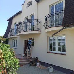 F.H.U Malors Marcin Słomski - Dwóch mężczyzn maluje elewację domu z balkonami, jeden na drabinie, drugi przy gruncie. Widoczny fragment ogrodu i kostka brukowa.