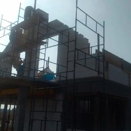 F.H.U Malors Marcin Słomski - Budowa domu z betonu komórkowego: murarze na rusztowaniu wznoszą ściany, widoczne szalunki stropowe, niebo w tle.