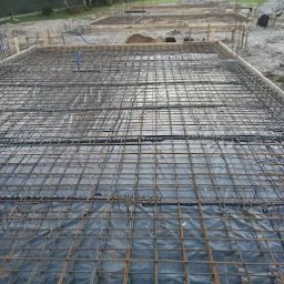 F.H.U Malors Marcin Słomski - Zbrojenie fundamentów pod budowę domu jednorodzinnego, widoczne szalunki i folia hydroizolacyjna. W tle usypana pryzma piasku.